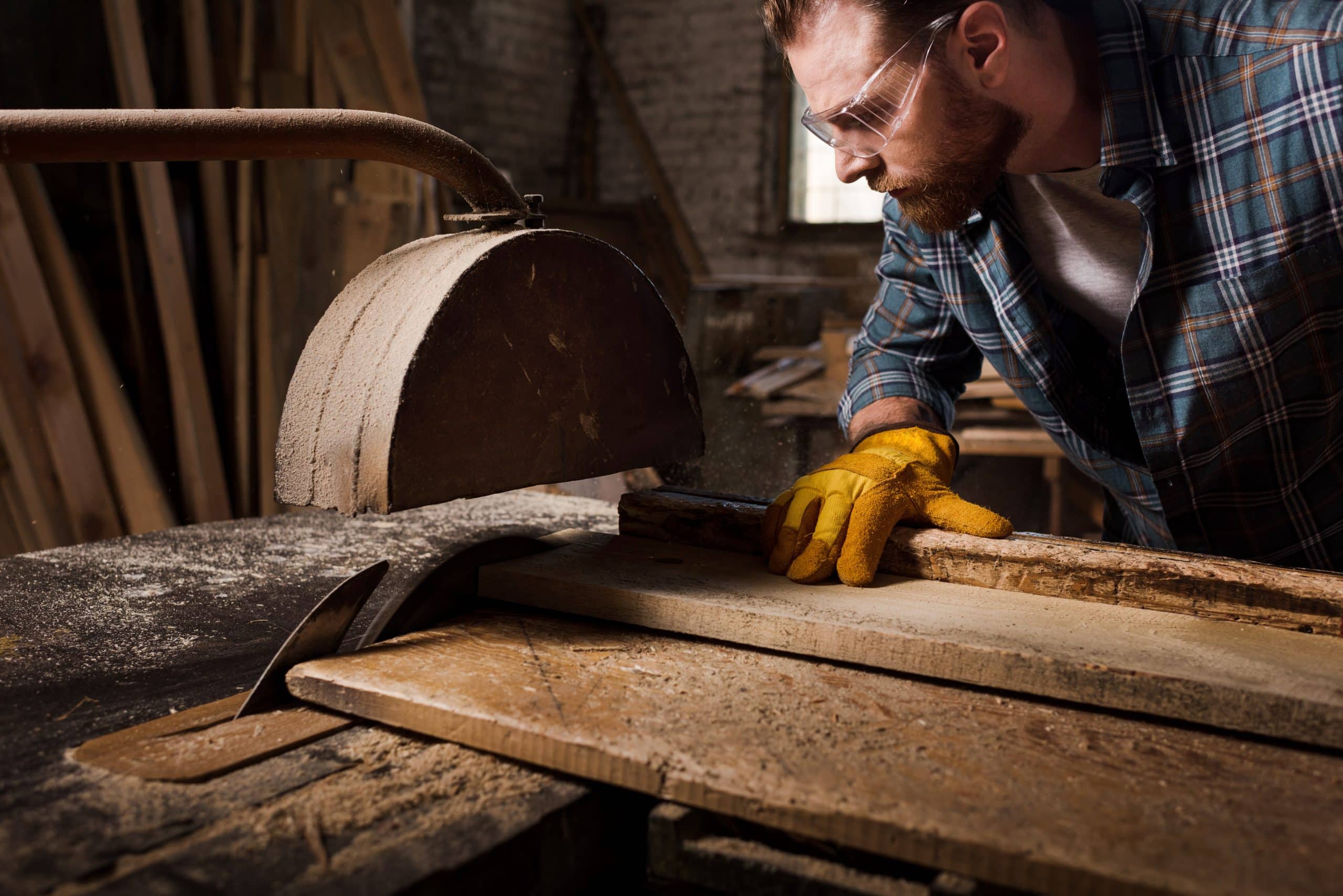 cropped shot of bearded carpenter in protective gloves and googles using machine saw at sawmill