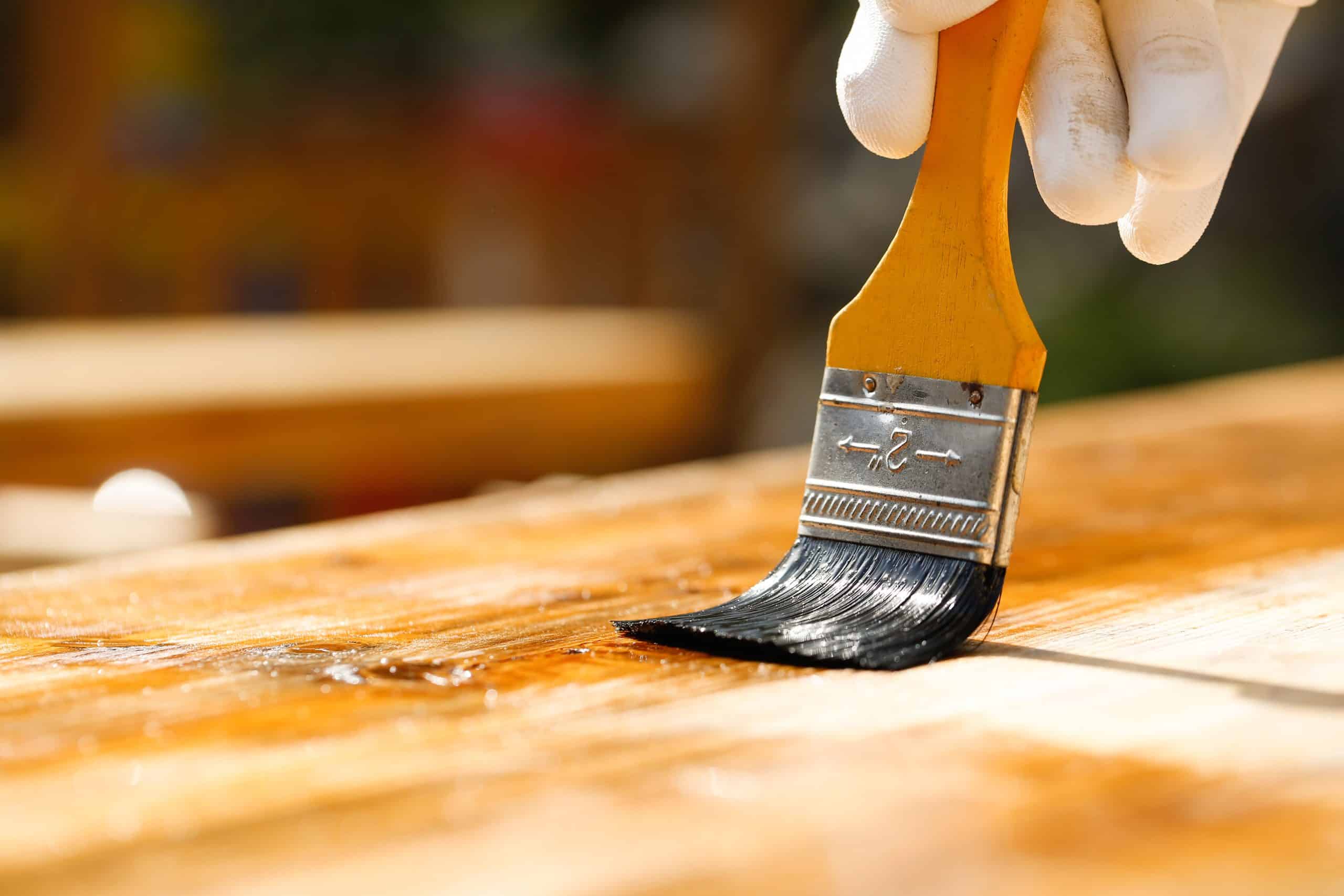 Gloved hand holding a paintbrush over wooden surface, protecting wood for exterior influences and weathering. Carpentry, wood treatment, hard at work, home improvement, do-it-yourself concept.