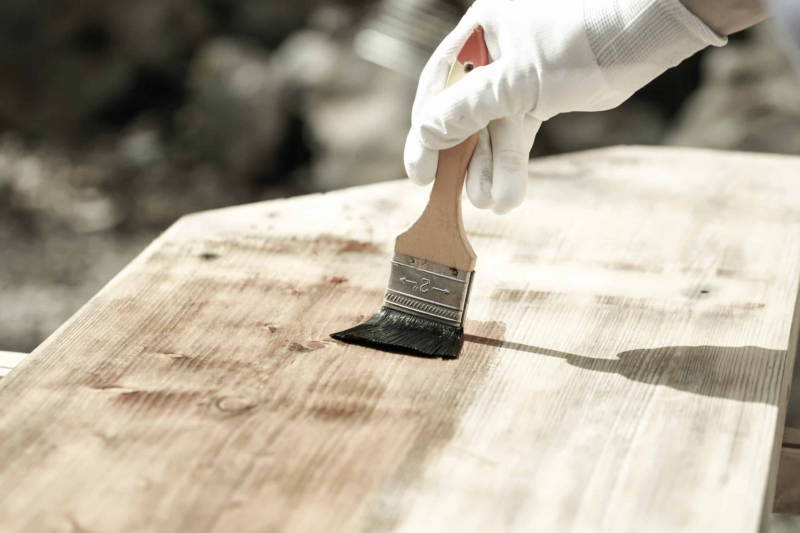 Gloved hand holding a paintbrush over wooden surface, protecting wood for exterior influences and weathering. Carpentry, wood treatment, hard at work, home improvement, do-it-yourself concept.